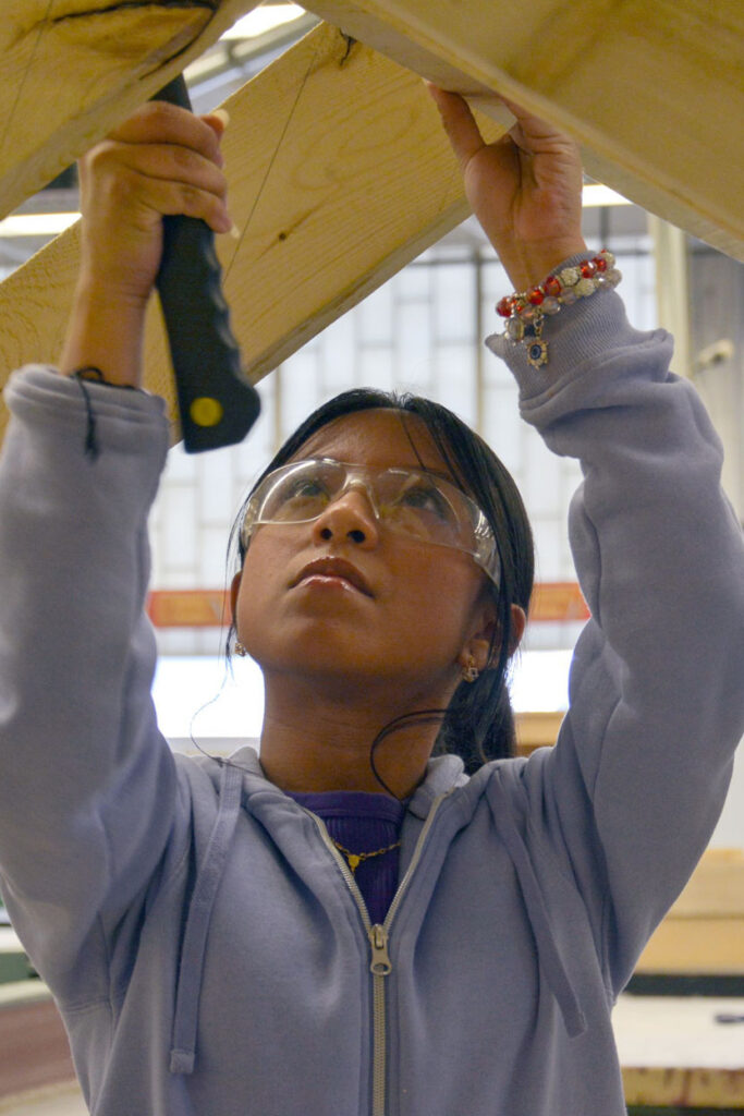 Student-Volunteers-Mission-of-Deeds | Mission of Deeds Young woman in safety goggles and a light purple hoodie using a screwdriver to fasten a wooden beam overhead in a workshop.